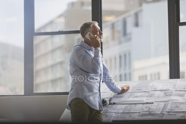 Architect talking on cell phone at blueprints in urban office — Stock Photo