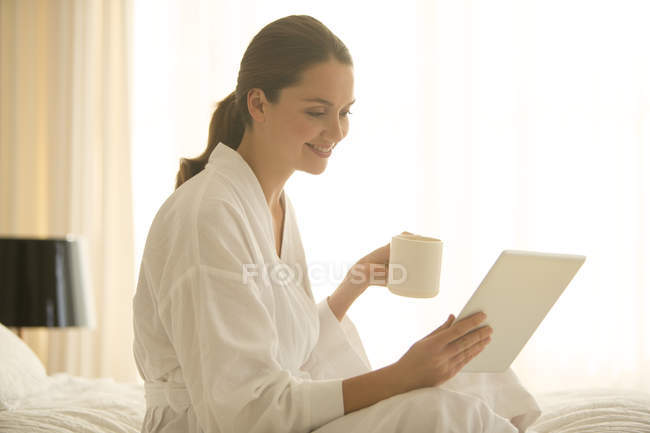 Жінка в халаті п'є каву і використовує цифровий планшет у спальні — стокове фото