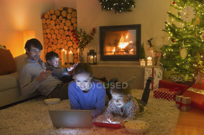 Familia relajante, el uso de ordenador portátil, tableta digital y teléfono celular en el ambiente sala de estar de Navidad - foto de stock