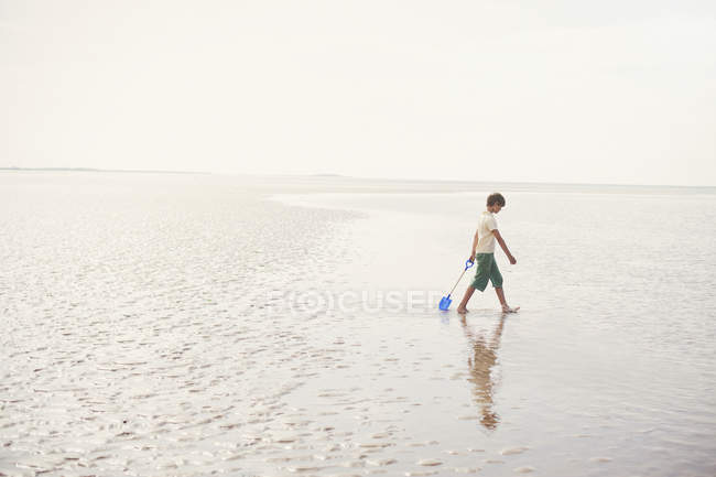 Menino andando com pá na areia molhada na praia de verão nublado — Fotografia de Stock