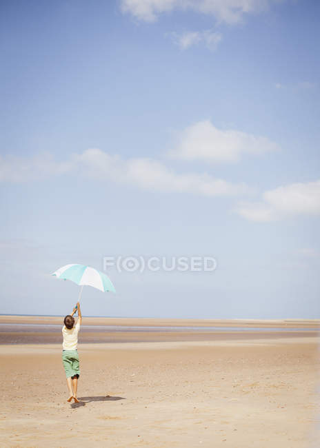 Мальчик держит полосатый зонтик над головой на солнечном летнем пляже под голубым небом — стоковое фото