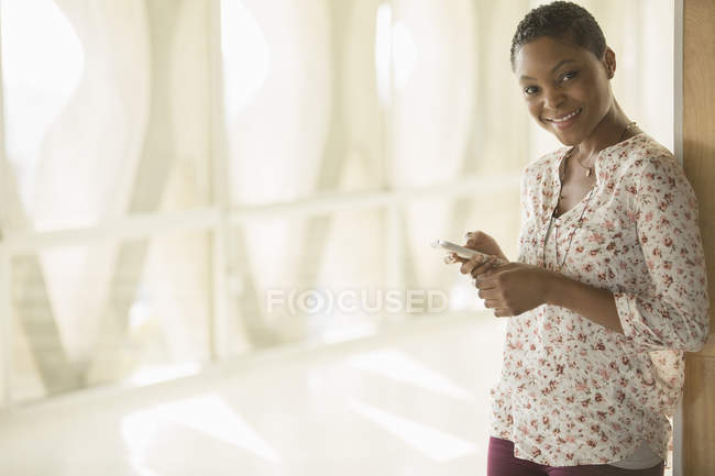 Портрет улыбающейся женщины, отправляющей смс с мобильного телефона в солнечном коридоре — стоковое фото