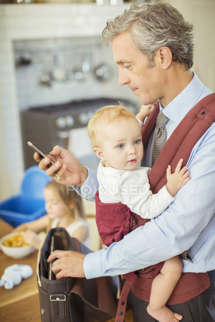 Padre sosteniendo al bebé y comprobando el teléfono celular - foto de stock