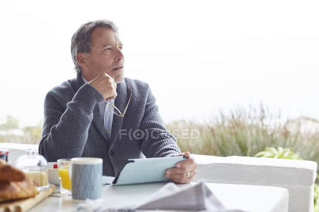 Uomo anziano pensieroso che utilizza tablet digitale a colazione sul patio — Foto stock
