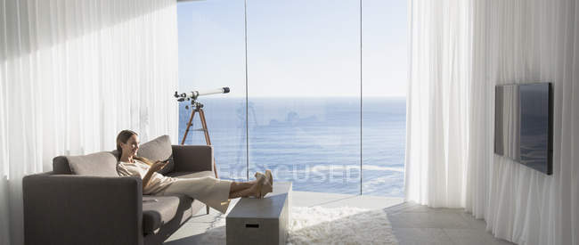 Mulher relaxante com os pés para cima, assistindo TV na moderna, casa de luxo vitrine interior sala de estar com vista para o mar ensolarado — Fotografia de Stock