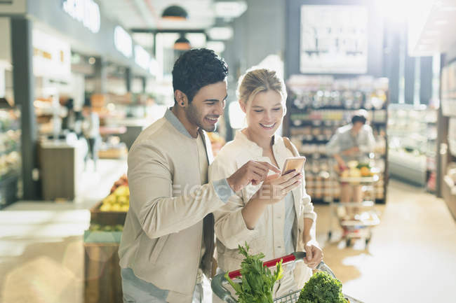 Jeune couple utilisant un téléphone cellulaire, épicerie dans le marché de l'épicerie — Photo de stock