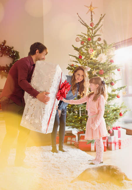 Padres dando un gran regalo de Navidad a su hija en la sala de estar junto al árbol de Navidad - foto de stock