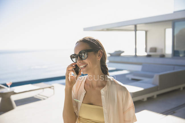 Усміхнена жінка розмовляє на смартфоні на сонячному сучасному, розкішному домашньому вітрині зовнішнього дворику — стокове фото