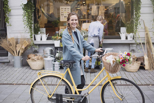 Retrato mujer sonriente caminando bicicleta con flores en cesta frente a la tienda exterior - foto de stock