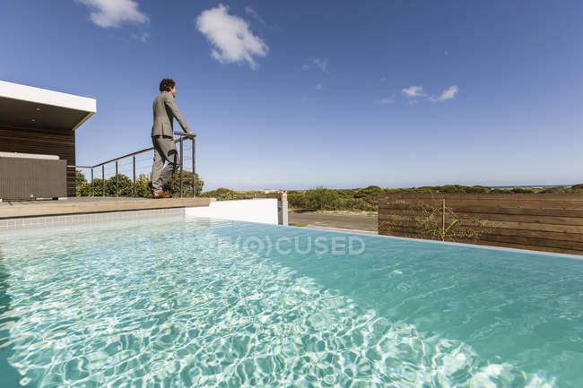 Бизнесмен, стоящий на роскошном солнечном патио с бесконечным бассейном, глядя на вид — стоковое фото