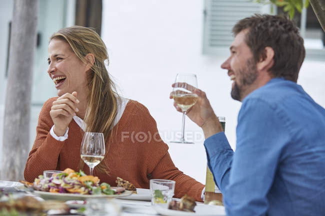 Riendo pareja bebiendo vino blanco y almorzando en la mesa del patio - foto de stock