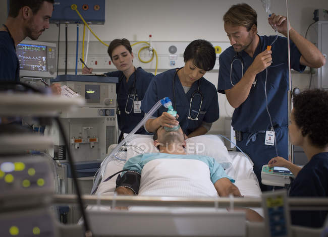 Médico sosteniendo máscara sobre la boca del paciente y médico controlando goteo IV en la unidad de cuidados intensivos - foto de stock