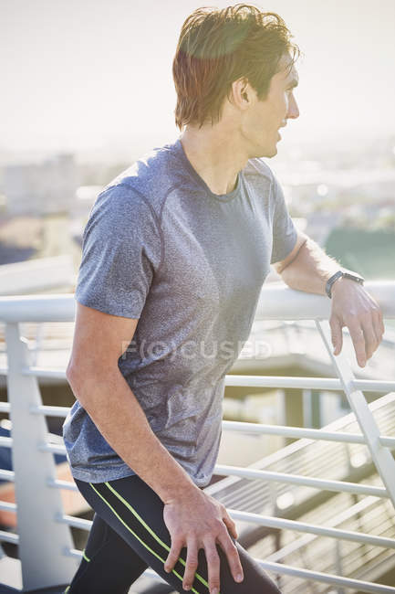 Suado masculino corredor descansando alongamento pernas no ensolarado urbano footbridge — Fotografia de Stock