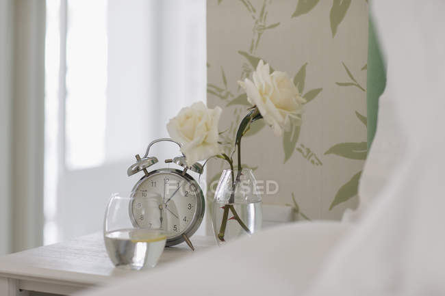 Rosa branca, despertador e vidro de água na mesa de cabeceira — Fotografia de Stock