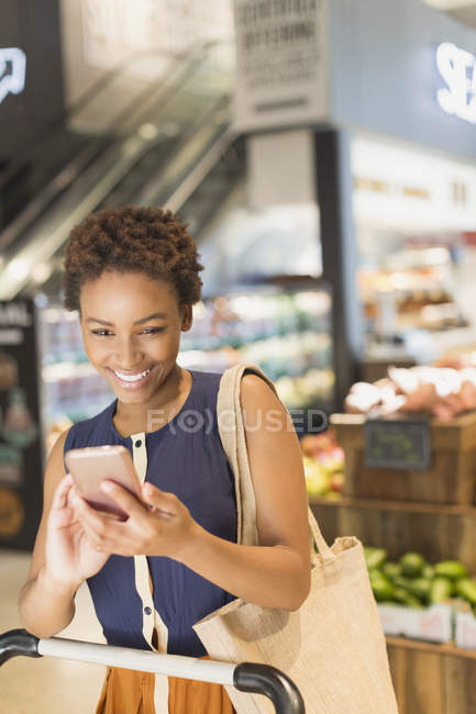 Улыбающаяся молодая женщина, использующая мобильный телефон на рынке продуктовых магазинов — стоковое фото