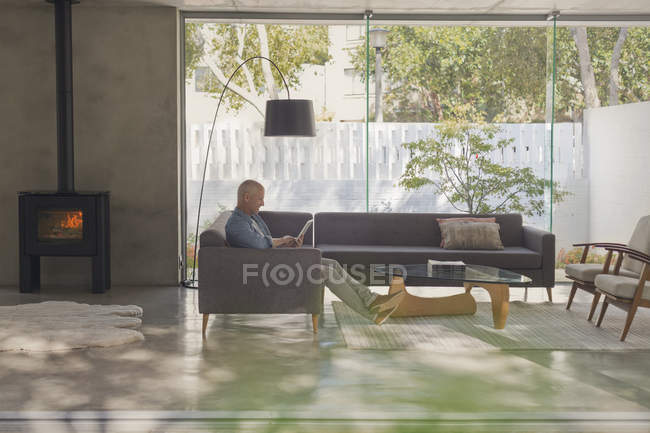 Homme mûr relaxant, utilisant tablette numérique sur canapé dans le salon avec cheminée — Photo de stock