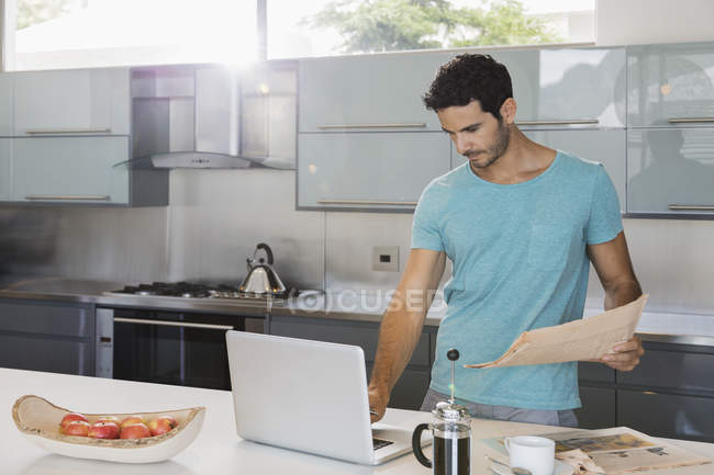 Hombre con periódico usando el ordenador portátil en la cocina - foto de stock