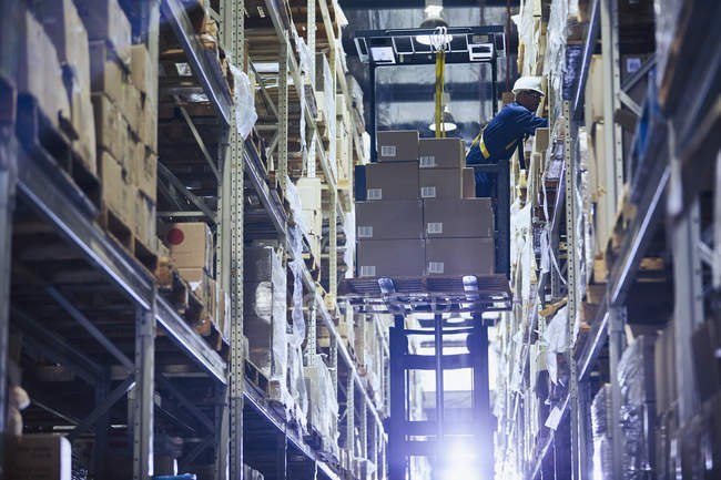 Travailleur fonctionnant chariot élévateur empilant des boîtes en carton sur les étagères de l'entrepôt de distribution — Photo de stock