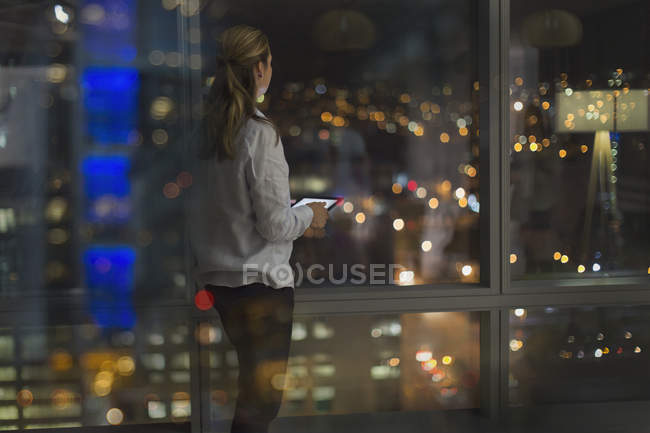 Pensive businesswoman with digital tablet working late, looking out urban office window at night — Stock Photo