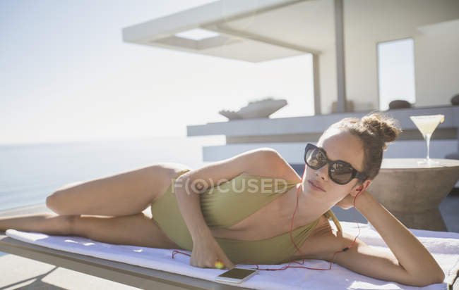 Retrato mulher confiante em roupa de banho banhos de sol no pátio de luxo ensolarado, ouvir música com telefone inteligente — Fotografia de Stock