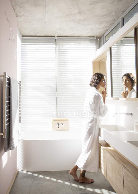 Frau im Bademantel untersucht Gesicht im Badezimmerspiegel — Stockfoto