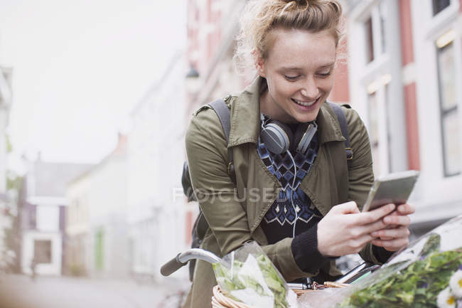 Sonriendo joven mujer mensajes de texto con el teléfono celular en bicicleta en la calle de la ciudad - foto de stock