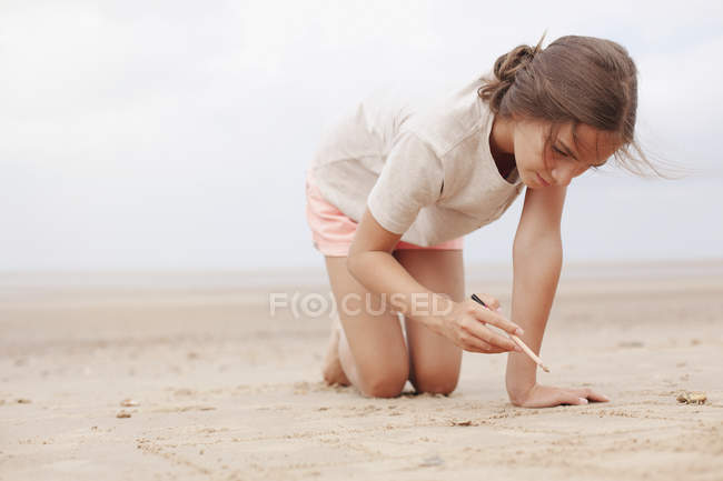 Chica con palo de escritura en la arena en la playa nublada de verano - foto de stock
