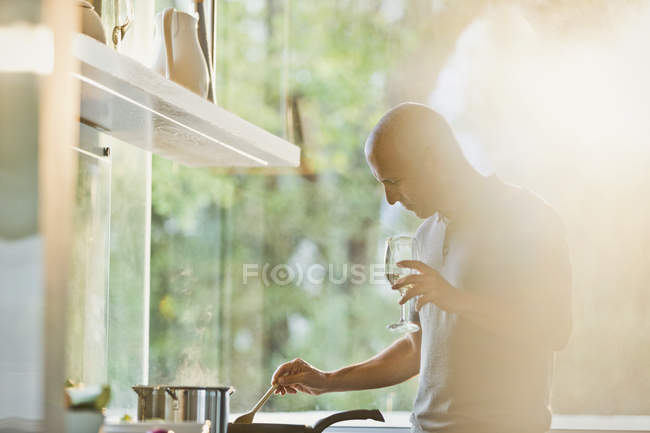 Mature man drinking white wine and cooking at stove in sunny kitchen — Stock Photo