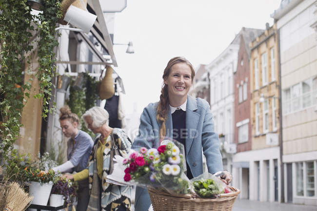 Donna sorridente in bicicletta con fiori nel cestino sulla strada della città — Foto stock