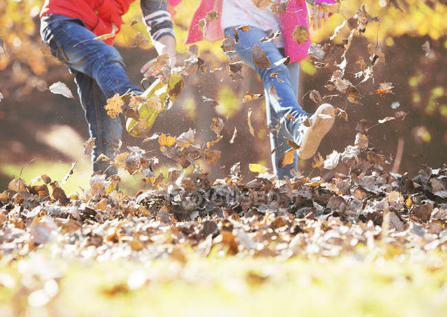 Imagen recortada de niño y niña pateando en hojas de otoño - foto de stock