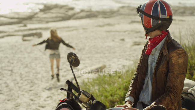Young man on motorcycle watching woman run onto beach — Stock Photo