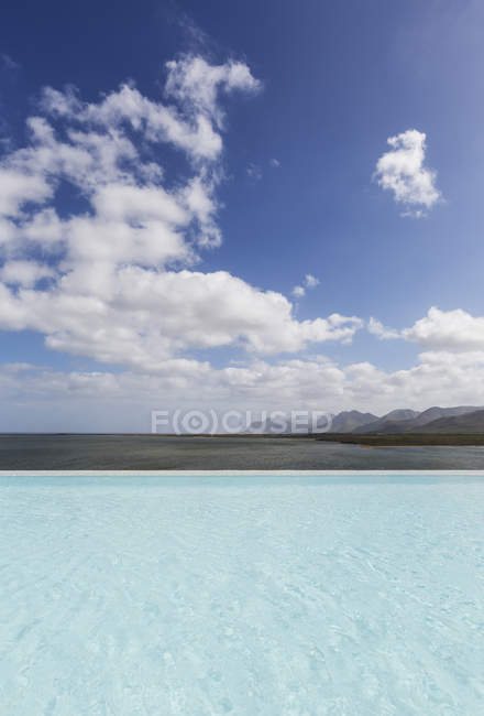 Sunny, tranquil infinity pool with ocean view under blue sky with clouds — Stock Photo