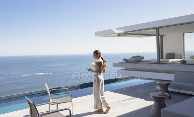 Donna a piedi e sms con smart phone sul soleggiato moderno, lussuoso patio vetrina casa con vista sull'oceano — Foto stock