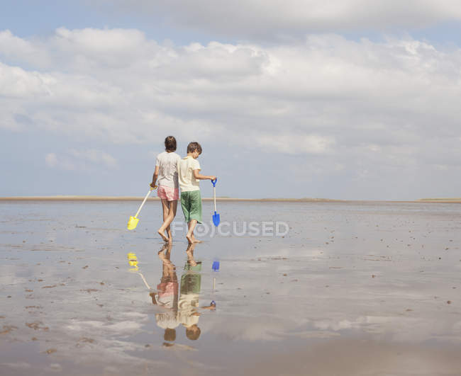 Brother and sister walking with shovels in wet sand on sunny summer beach — Stock Photo