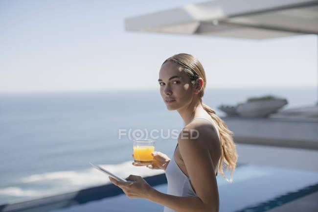 Mujer retrato usando tableta digital y beber jugo de naranja en patio de lujo soleado con vista al mar - foto de stock