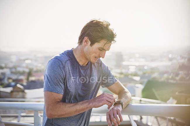 Потный бегун-мужчина отдыхает, проверяя умные часы фитнес-трекер на солнечных городских перилах — стоковое фото