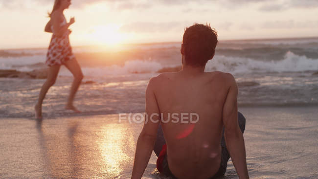 Young man watching woman running in ocean surf at sunset — Stock Photo