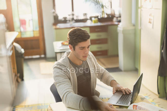 Joven estudiante universitario estudiando en el portátil en la cocina - foto de stock