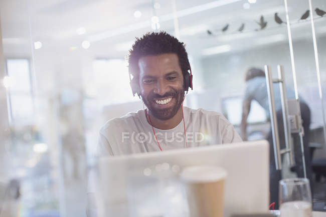 Улыбающийся, уверенный в себе креативный бизнесмен с наушниками с помощью ноутбука в офисе — стоковое фото