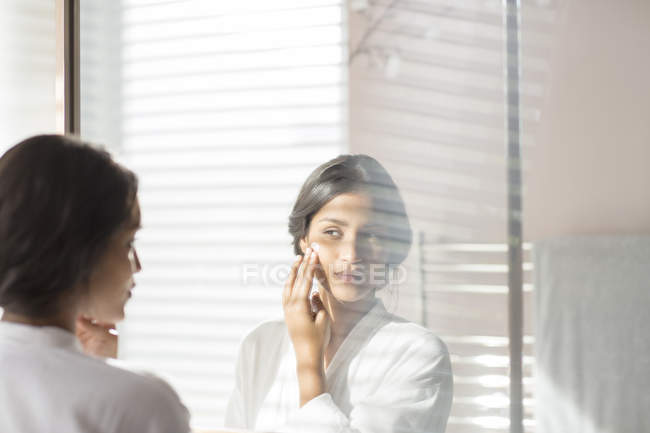 Femme appliquant une crème hydratante sur la joue dans le miroir de salle de bain — Photo de stock
