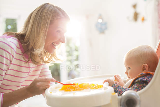 Rubia madre alimentación bebé niña en silla alta - foto de stock