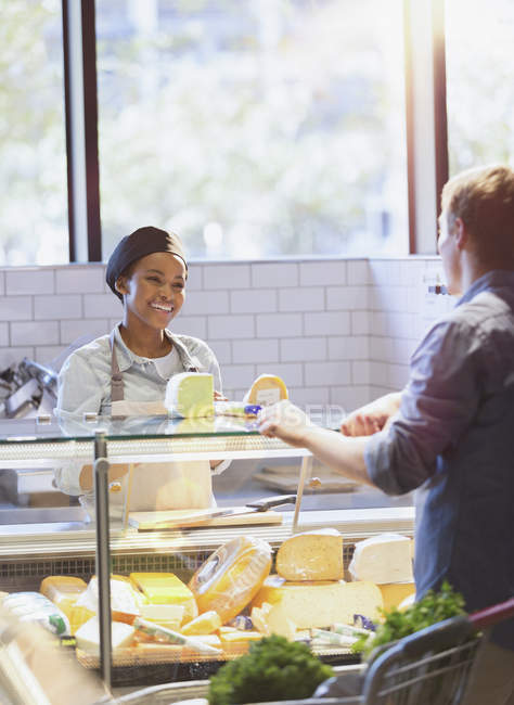 Smiling young woman helping customer at cheese counter in grocery store market — Stock Photo