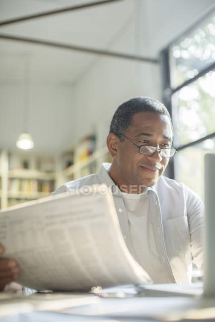 Hombre mayor leyendo el periódico y utilizando el ordenador portátil - foto de stock