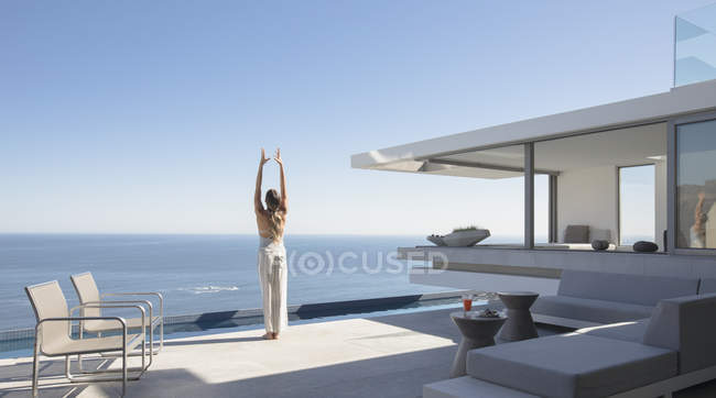 Donna che pratica yoga montagna posa sul soleggiato moderno, casa di lusso vetrina patio esterno con vista sull'oceano — Foto stock