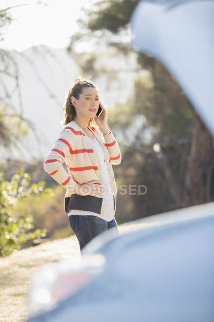 Mujer hablando por teléfono celular con capucha de automóvil levantada al borde de la carretera - foto de stock