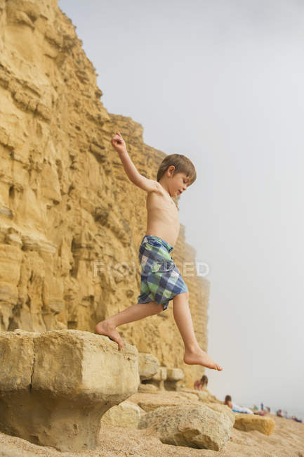 Menino em calções de banho pulando na praia rock — Fotografia de Stock