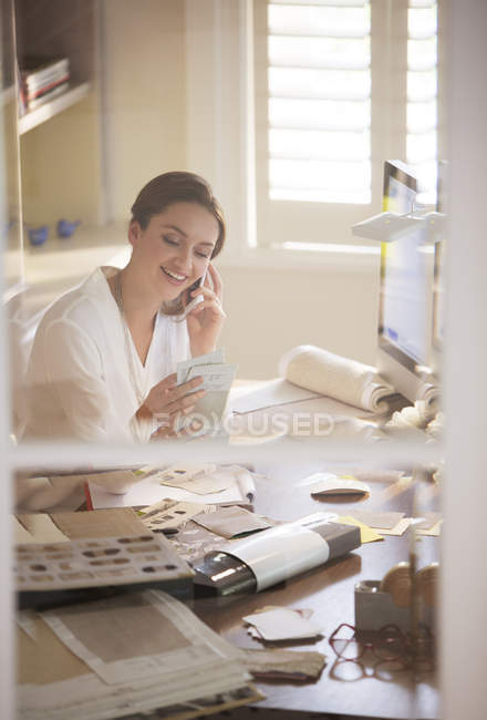 Designer de interiores examinando amostras e falando no telefone celular no escritório em casa — Fotografia de Stock