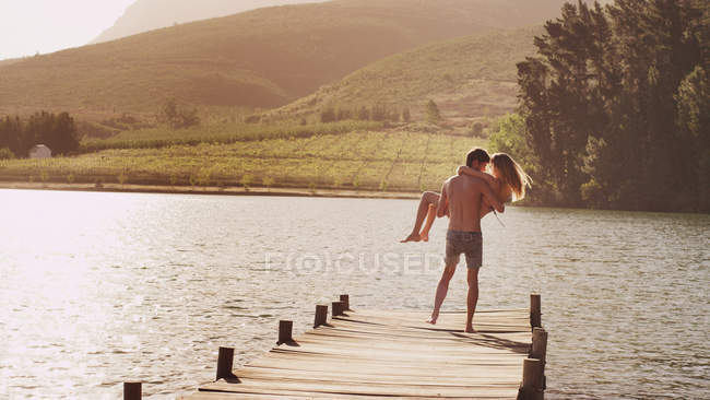 Hombre joven llevando a la mujer en el muelle soleado junto al lago - foto de stock