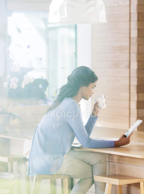 Frau trinkt Kaffee und benutzt digitales Tablet an der Frühstückstheke — Stockfoto