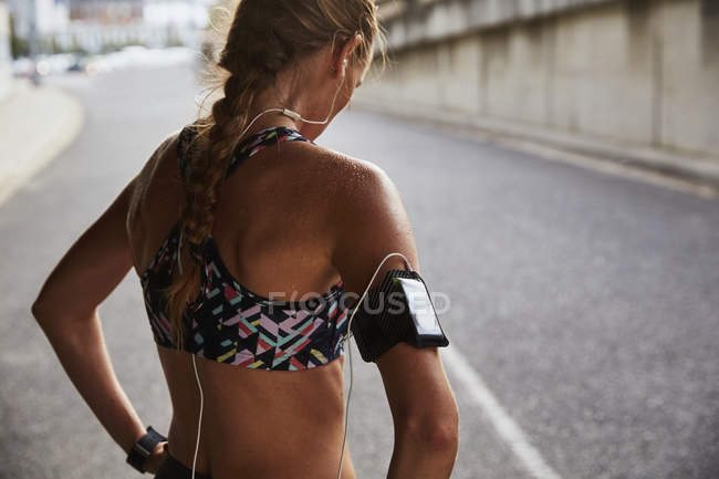 Fit female runner in sports bra with mp3 player armband and headphones resting on urban street — Stock Photo